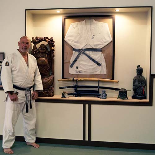 Jan Rust in judogi, in de dojo van Sportcentrum Rust, naast zijn ingelijste judogi en zwarte band